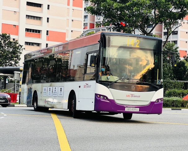 A public transit bus in motion on an urban road. The bus has a purple and white color scheme with the SBS Transit logo. It is displaying route number 142 and is surrounded by high-rise residential buildings and greenery.