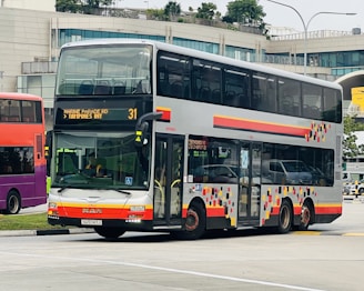 A double-decker bus is parked on a road near a modern building with large windows. The bus has a colorful geometric pattern on its side and digital displays indicating its route to 'Marine Parade Rd' and 'Tampines Int'. Several vehicles are visible in the background, suggesting a bustling urban environment.