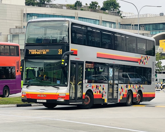 A double-decker bus is parked on a road near a modern building with large windows. The bus has a colorful geometric pattern on its side and digital displays indicating its route to 'Marine Parade Rd' and 'Tampines Int'. Several vehicles are visible in the background, suggesting a bustling urban environment.