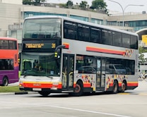A double-decker bus is parked on a road near a modern building with large windows. The bus has a colorful geometric pattern on its side and digital displays indicating its route to 'Marine Parade Rd' and 'Tampines Int'. Several vehicles are visible in the background, suggesting a bustling urban environment.