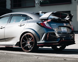A sleek, modern Honda Civic is parked in a lot, showcasing its sporty design. The car features a prominent rear wing and glossy black alloy wheels with red accents. The sunlight reflects off its metallic gray body, highlighting its aerodynamic shape.