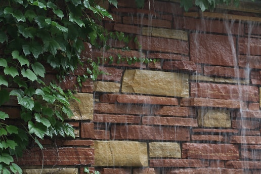 A stone wall with irregularly shaped bricks in shades of brown and beige, partially covered with lush green ivy. Water streams gently down the surface of the wall, creating a serene waterfall effect.