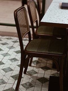 Two wooden chairs with woven backrests and cushioned seats are positioned next to a terrazzo-topped table. The floor features a tile pattern with green and white diamond shapes.
