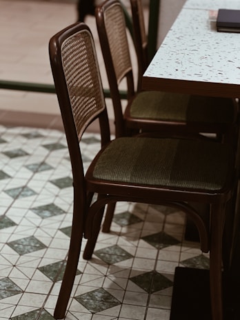 Two wooden chairs with woven backrests and cushioned seats are positioned next to a terrazzo-topped table. The floor features a tile pattern with green and white diamond shapes.
