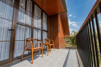 A modern balcony with wooden paneling is equipped with two wooden chairs. The glass doors are adorned with sheer white curtains, and the floor is covered with light gray tiles. The railing features black vertical bars, and the sky is a clear blue with a few scattered clouds, with some greenery visible in the distance.