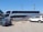 A large double-decker bus with 'Ejecutivo Premium' written on its side is parked near a fence in a parking lot. The bus is white with blue and red stripes. Two cars, one green and one white, are parked nearby. The scene is set on a clear day with a blue sky, power lines, and part of a building visible in the background.