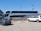 A large double-decker bus with 'Ejecutivo Premium' written on its side is parked near a fence in a parking lot. The bus is white with blue and red stripes. Two cars, one green and one white, are parked nearby. The scene is set on a clear day with a blue sky, power lines, and part of a building visible in the background.