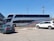 A large double-decker bus with 'Ejecutivo Premium' written on its side is parked near a fence in a parking lot. The bus is white with blue and red stripes. Two cars, one green and one white, are parked nearby. The scene is set on a clear day with a blue sky, power lines, and part of a building visible in the background.