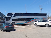 A large double-decker bus with 'Ejecutivo Premium' written on its side is parked near a fence in a parking lot. The bus is white with blue and red stripes. Two cars, one green and one white, are parked nearby. The scene is set on a clear day with a blue sky, power lines, and part of a building visible in the background.