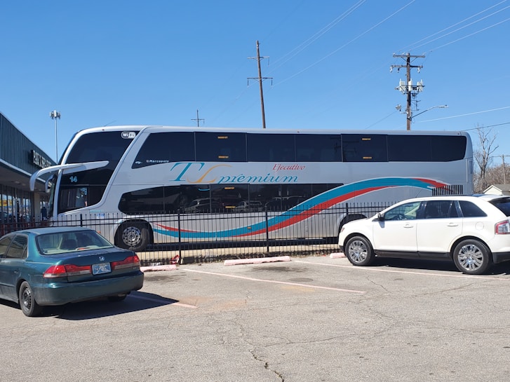 A large double-decker bus with 'Ejecutivo Premium' written on its side is parked near a fence in a parking lot. The bus is white with blue and red stripes. Two cars, one green and one white, are parked nearby. The scene is set on a clear day with a blue sky, power lines, and part of a building visible in the background.