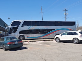 A large double-decker bus with 'Ejecutivo Premium' written on its side is parked near a fence in a parking lot. The bus is white with blue and red stripes. Two cars, one green and one white, are parked nearby. The scene is set on a clear day with a blue sky, power lines, and part of a building visible in the background.