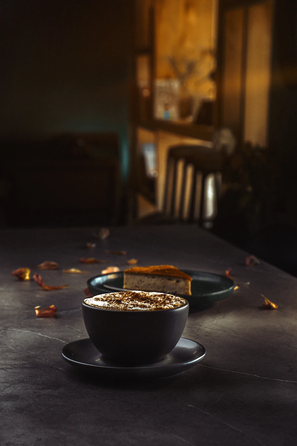 The image shows a black bowl filled with a warm beverage, possibly coffee or tea, placed on a dark gray table. The bowl is positioned centrally and slightly tilted to the left side. To the right of the bowl, there's another small black bowl containing a slice of cake. Both bowls are set against a blurred background that appears to be a kitchen countertop with various items scattered around it.