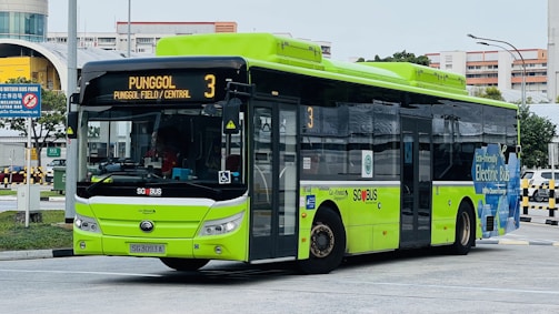 A green and black electric city bus with signage indicating it is on route 3 to Punggol Field/Central. The bus displays markings suggesting it is an eco-friendly electric bus operated by SBS Transit. It is stationed at a bus interchange area with buildings and road signs in the background.