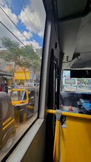 A view from inside a bus, looking out through the window. On the street, there are several auto-rickshaws and a truck in the background. The outside scene shows a city environment with trees, buildings, and signboards under a partly cloudy sky.