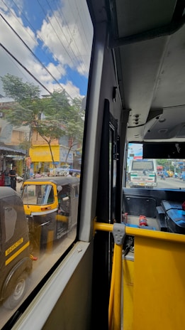 A view from inside a bus, looking out through the window. On the street, there are several auto-rickshaws and a truck in the background. The outside scene shows a city environment with trees, buildings, and signboards under a partly cloudy sky.
