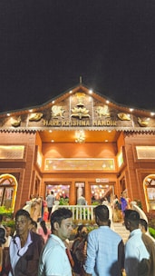 A temple entrance with the name Hare Krishna Mandir prominently displayed. The building is adorned with intricate carvings and illuminated at night, creating a festive atmosphere. A number of people are gathered at the entrance, some climbing the steps and others standing in groups, indicating a social or religious gathering.