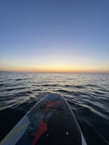 A paddleboard floats on a vast expanse of calm ocean, with gentle waves. The sky is a gradient of soft blues, transitioning into a warm yellow and orange glow at the horizon, indicating either sunrise or sunset. Light reflects off the water's surface, highlighting the serene atmosphere.
