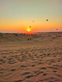 A serene desert landscape during sunset with vibrant hues in the sky. The sun is low on the horizon, emitting a warm glow. Several people are gathered in the distance, some engaging in parasailing. The sand is patterned with footprints and ripples, displaying a golden tone.