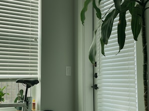 White venetian blinds cover a large window, allowing a diffused light to enter the room. A tall green plant stands beside the window, its leaves reaching towards the blinds. A bicycle seat is visible in the lower left corner, next to a small container of oil or glue. The ambiance is calm and tidy.