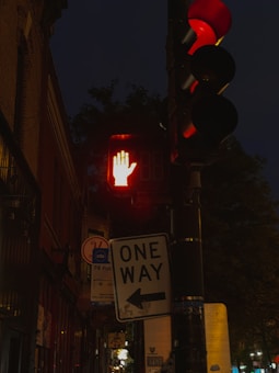A traffic signal at night, featuring illuminated red lights for vehicles and a red hand pedestrian signal indicating not to walk. There are various signs on the pole, including a one-way street sign with an arrow pointing left and a bus stop sign in the background. The surroundings include buildings and trees in the dark.