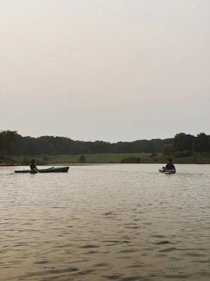 Two kayaks gliding smoothly on a clear lake framed by tall pine trees under a bright sky.