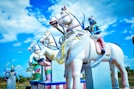 Statues of warriors are riding white horses with a vivid blue sky and fluffy clouds in the background. The warriors are painted in a combination of blue, pink, and gold, with detailed clothing and weapons. The horses are adorned with colorful decorations and sashes.