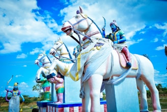 Statues of warriors are riding white horses with a vivid blue sky and fluffy clouds in the background. The warriors are painted in a combination of blue, pink, and gold, with detailed clothing and weapons. The horses are adorned with colorful decorations and sashes.