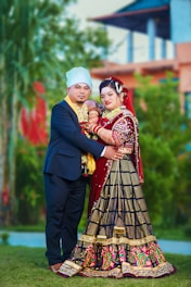 A couple dressed in traditional attire poses together in an outdoor setting. The woman is wearing an elaborate red and black dress with intricate embroidery, and the man is in a formal dark suit with a light-colored turban. They are standing close, surrounded by greenery and blurred architectural elements in the background.
