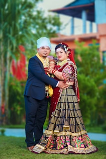 A couple dressed in traditional attire poses together in an outdoor setting. The woman is wearing an elaborate red and black dress with intricate embroidery, and the man is in a formal dark suit with a light-colored turban. They are standing close, surrounded by greenery and blurred architectural elements in the background.