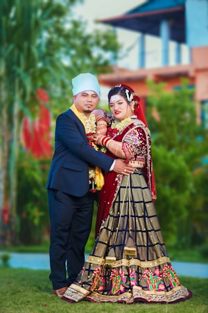 A couple dressed in traditional attire poses together in an outdoor setting. The woman is wearing an elaborate red and black dress with intricate embroidery, and the man is in a formal dark suit with a light-colored turban. They are standing close, surrounded by greenery and blurred architectural elements in the background.