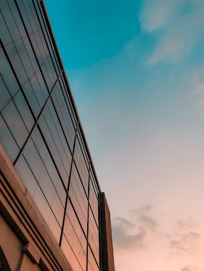 A modern building with a glass facade reflecting the sky during sunset. The sky displays hues of blue and orange, creating a serene backdrop. The architectural lines of the building lead the eye upwards.