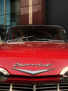 A vintage red Chevrolet car is displayed, featuring its emblem prominently on the hood. The car's front grill and windshield are visible, with reflections of nearby buildings on the glass.