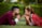 A couple is lying on the grass face-to-face, gazing into each other's eyes. The woman is dressed in a traditional red and white outfit with gold jewelry, while the man is wearing a maroon suit. They both have their hands clasped under their chins, creating a serene and intimate moment.