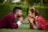 A couple is lying on the grass face-to-face, gazing into each other's eyes. The woman is dressed in a traditional red and white outfit with gold jewelry, while the man is wearing a maroon suit. They both have their hands clasped under their chins, creating a serene and intimate moment.