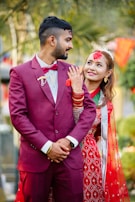 A couple is dressed in traditional attire, likely for a wedding ceremony. The man is wearing a maroon suit with a bow tie, while the woman is in a red and green saree adorned with jewelry and mehndi on her hand. They both appear to be smiling and looking content, with lush greenery and festive decorations in the background.