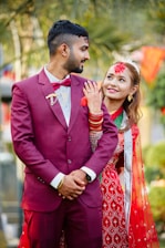 A couple is dressed in traditional attire, likely for a wedding ceremony. The man is wearing a maroon suit with a bow tie, while the woman is in a red and green saree adorned with jewelry and mehndi on her hand. They both appear to be smiling and looking content, with lush greenery and festive decorations in the background.