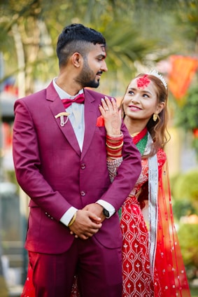 A couple is dressed in traditional attire, likely for a wedding ceremony. The man is wearing a maroon suit with a bow tie, while the woman is in a red and green saree adorned with jewelry and mehndi on her hand. They both appear to be smiling and looking content, with lush greenery and festive decorations in the background.