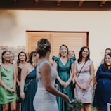 A group of women, dressed in elegant attire, stands together enjoying a moment of joy and laughter. The central figure, possibly a bride, has her back to the camera, and the women in the foreground are facing her with smiles. The setting appears to be outdoors with a building featuring wooden doors in the background.