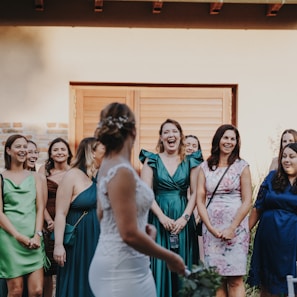 A group of women, dressed in elegant attire, stands together enjoying a moment of joy and laughter. The central figure, possibly a bride, has her back to the camera, and the women in the foreground are facing her with smiles. The setting appears to be outdoors with a building featuring wooden doors in the background.