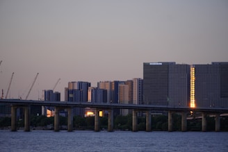 A bridge stretches over a calm body of water with modern high-rise buildings in the background. Several construction cranes are positioned in front of the buildings, indicating ongoing development. The sky is clear with a hint of dusk, and a warm, reflective light is visible on one of the buildings.