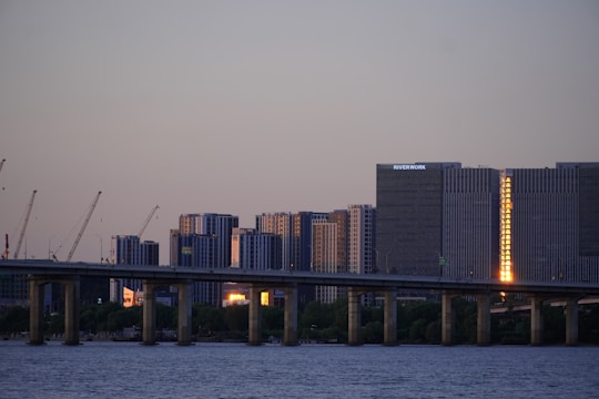 A bridge stretches over a calm body of water with modern high-rise buildings in the background. Several construction cranes are positioned in front of the buildings, indicating ongoing development. The sky is clear with a hint of dusk, and a warm, reflective light is visible on one of the buildings.