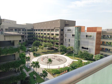 A modern courtyard surrounded by multi-story buildings with a lot of greenery on balconies and in the garden area. The courtyard features a circular pathway with palm trees and benches. The architecture is contemporary with a mix of gray concrete and wooden panels.