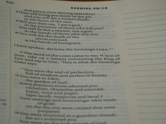 A close-up of a Bible page showing text from Ezekiel 28:8-14. The text includes references to a shining splendor, a violent death, and descriptions of Eden with precious stones.