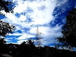 A high voltage transmission tower stands amidst a backdrop of vibrant blue sky and scattered white clouds. Surrounding the tower are silhouettes of leafy trees, providing a natural contrast to the industrial structure.