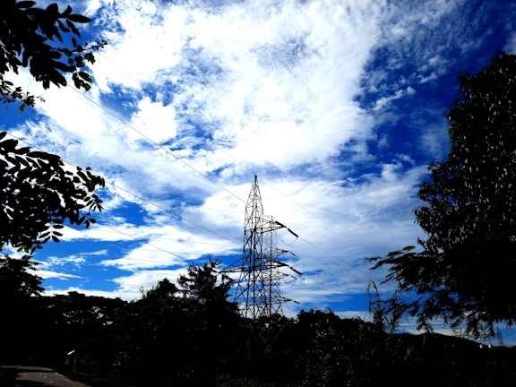 A high voltage transmission tower stands amidst a backdrop of vibrant blue sky and scattered white clouds. Surrounding the tower are silhouettes of leafy trees, providing a natural contrast to the industrial structure.