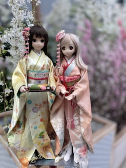 Two dolls dressed in traditional Japanese kimonos are positioned in a serene, floral setting. The dolls have large eyes and elaborately styled hair with floral accessories. One doll wears a light yellow kimono with intricate patterns and holds a tray with colorful sweets. The other doll is in a soft pink kimono adorned with floral designs.