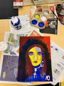 A painted portrait of a woman with bold, contrasting colors lies on a table surrounded by art supplies. The woman has long hair and is adorned with large hoop earrings and a cross necklace. Various paint bottles, a palette with several colors, and scattered magazines or newspapers are visible on the table.