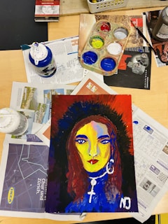 A painted portrait of a woman with bold, contrasting colors lies on a table surrounded by art supplies. The woman has long hair and is adorned with large hoop earrings and a cross necklace. Various paint bottles, a palette with several colors, and scattered magazines or newspapers are visible on the table.