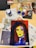 A painted portrait of a woman with bold, contrasting colors lies on a table surrounded by art supplies. The woman has long hair and is adorned with large hoop earrings and a cross necklace. Various paint bottles, a palette with several colors, and scattered magazines or newspapers are visible on the table.