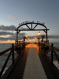 A wooden pier extends into the sea with warm street lamps illuminating its path. The sky is a mix of dark clouds and subtle sunset hues. The entrance is marked by a sign reading 'Marbella Club' and features palm trees lining the walkway.